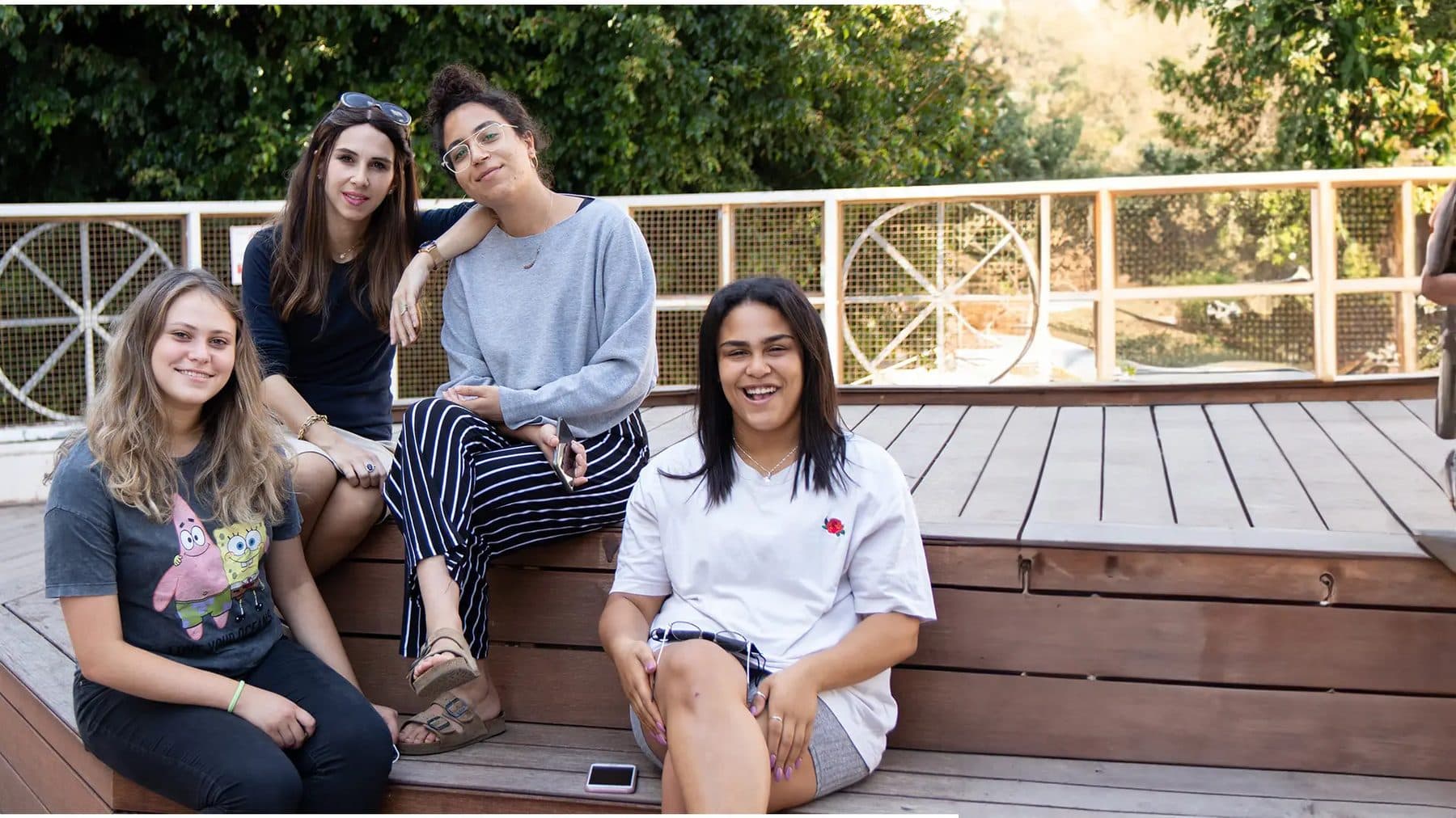 בנות שירות של שילה יושבות יחד על דק עץ, צוחקות